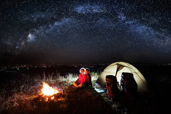 Acampamento nocturno perto da cidade. Casal jovem tendo um resto perto de fogueira e tenda com mochilas, sob céu noturno bonito cheio de estrelas. Exposição longa — Fotografia de Stock