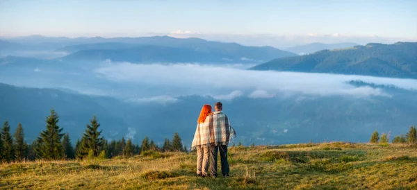 Romantische paar bedekt met een deken staande op een heuvel geniet van een ochtend nevel over de bergen. Achteraanzicht. Panorama — Stockfoto