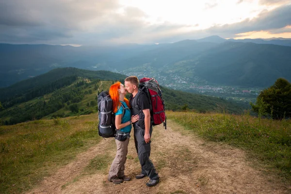 Mutlu çift yürüyüşçü dağlarda yolda öpüşme. Arka plan - yüce dağları, ormanları, tepeleri ve bulutlar gökyüzü — Stok fotoğraf