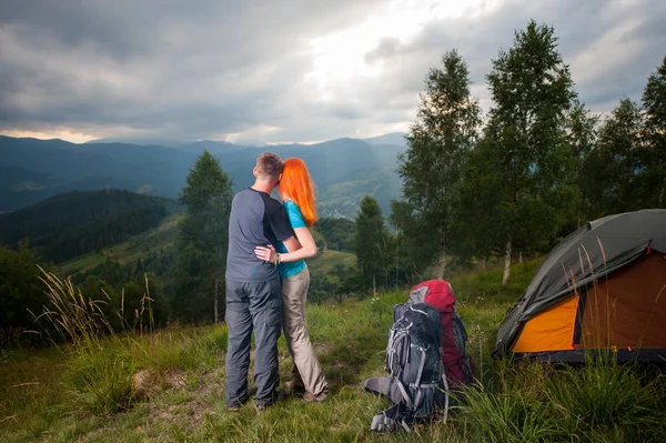 Abrazando a la pareja de pie cerca del campamento y mirando a la distancia en las montañas, bosques y golpeando los rayos del sol a través de un cielo nublado al atardecer — Foto de Stock