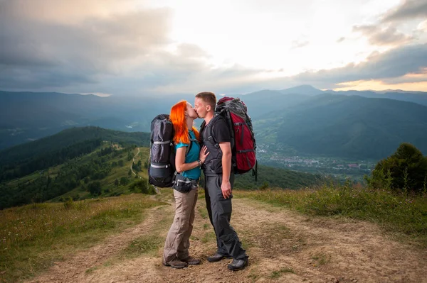 Çift backpackers sevgi dolu dağlarda yolda öpüyor. Arka plan - yüce dağları, ormanları, tepeleri ve bulutlar gökyüzü — Stok fotoğraf