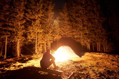 Kamp ateşi ve çadır yakınında oturan adam