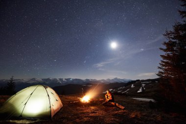 Kamp ateşi ve çadır yakınında oturan adam