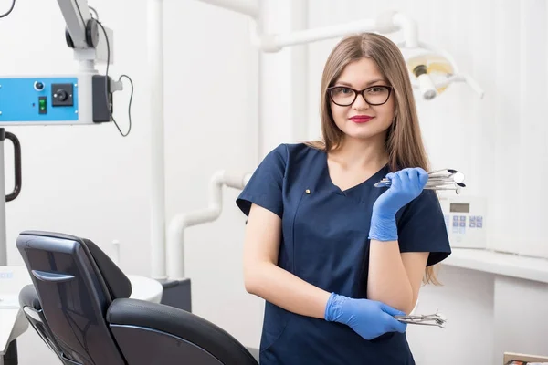 Πορτραίτο του νέους ελκυστικές οδοντίατρος κρατώντας οδοντιατρικές συσκευές στο σύγχρονο οδοντιατρείο. Γιατρός φοράει γυαλιά, μπλε στολή, μπλε γάντια και ψάχνει για την κάμερα. Οδοντιατρική — Φωτογραφία Αρχείου