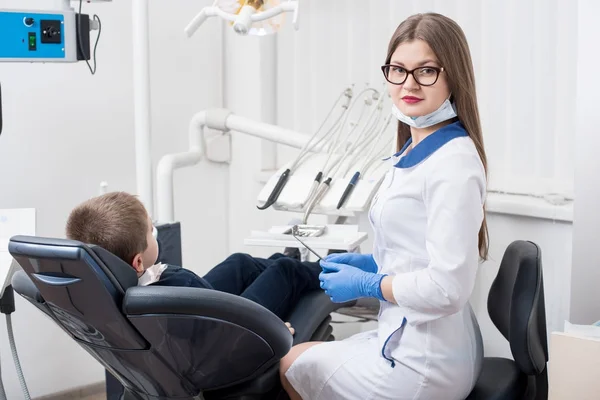 Female dentists examining and working with boy patient. Kid boy in dentist's chair. Doctor looks at camera. Dentist's office