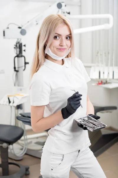 Retrato de una joven y hermosa dentista sosteniendo una herramienta dental en el consultorio dental de Morden. Doctor con guantes negros y mirando a la cámara. Odontología — Foto de Stock