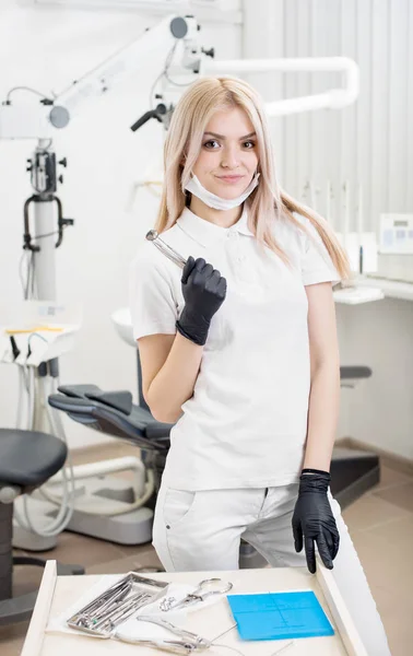 Porträt einer jungen attraktiven Zahnärztin, die zahnärztliche Geräte in der morden Zahnarztpraxis hält. Arzt mit Maske, schwarzen Handschuhen und Blick in die Kamera. Zahnmedizin — Stockfoto