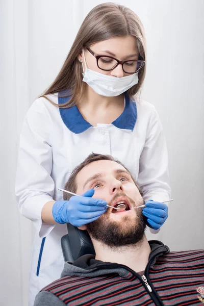 Jovem dentista segurando ferramentas dentárias espelho e sonda e fazendo primeiro check-up paciente masculino no consultório odontológico. Médico vestindo uniforme branco, óculos, máscara e luvas azuis. Odontologia — Fotografia de Stock