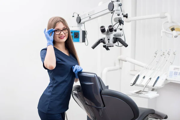 Πορτρέτο της ευτυχισμένη γυναίκα βοηθός στο οδοντικό γραφείο morden. Γιατρός φοράει γυαλιά, μπλε στολή, μπλε γάντια και ψάχνει για την κάμερα. Οδοντιατρική — Φωτογραφία Αρχείου