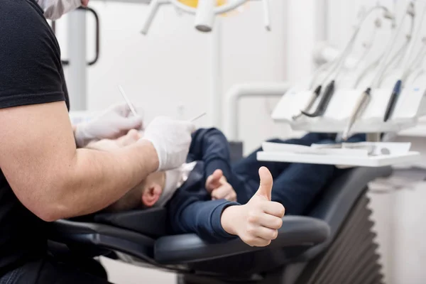 Pediatric tandläkare undersöker tänder av pojken patient i tandvårdsklinik med dental verktyg. Patienten visar tummen. Fokusera på patientens hand. Tandvård — Stockfoto