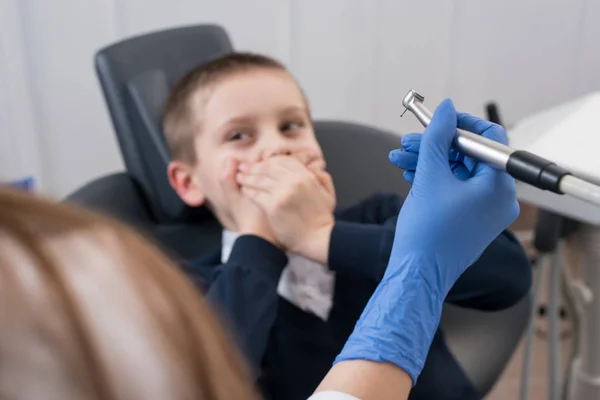 Dentysta w rękawice, trzymając w ręku dentystyczny wiertło i dzieciak przestraszony przez dentystów obejmuje jej usta. Ręka z wiertła stomatologiczne jest w centrum uwagi — Zdjęcie stockowe