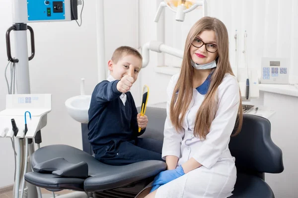 Όμορφο παιδί αγόρι χαμογελά στην καρέκλα του οδοντιάτρου και δείχνοντας μπράβο χειρονομίες καλής κατηγορίας. Ο γιατρός και το παιδί κοιτάζει κάμερα. — Φωτογραφία Αρχείου