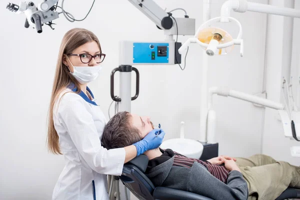 Νεαρή γυναίκα οδοντίατρος με μάσκα κρατώντας ΟΔΟΝΤΙΑΤΡΙΚΑ ΕΡΓΑΛΕΙΑ - καθρέφτη και καθετήρα και ετοιμάζεται για την οδοντιατρική διαδικασία με Αρσενικός ασθενής στο οδοντιατρείο. Γιατρό που ψάχνετε στην κάμερα. Οδοντιατρική — Φωτογραφία Αρχείου
