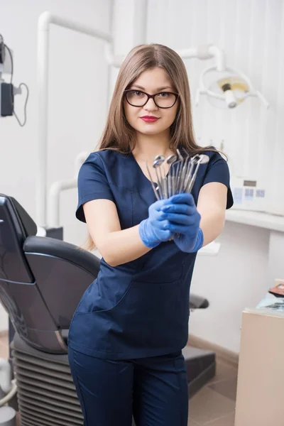 Πορτραίτο του ελκυστική γυναίκα οδοντίατρος κρατώντας ΟΔΟΝΤΙΑΤΡΙΚΑ ΕΡΓΑΛΕΙΑ στο οδοντικό γραφείο morden. Γιατρός φοράει γυαλιά, μπλε στολή, μπλε γάντια και ψάχνει για την κάμερα. Οδοντιατρική — Φωτογραφία Αρχείου