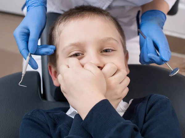 El primer plano del chico se cubre la boca en la silla del dentista y mira a la cámara. Las manos del médico en guantes y mantiene las herramientas — Foto de Stock