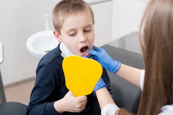 Niño en silla de dentista se mira en el espejo y el médico en guantes azules le muestra sus dientes . —  Fotos de Stock