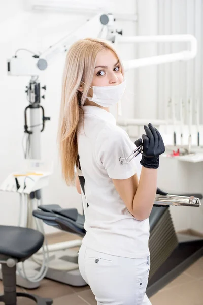 Porträt einer jungen Zahnärztin, die zahnärztliche Geräte in der morden Zahnarztpraxis hält. Arzt mit Maske, schwarzen Handschuhen und Blick in die Kamera. Zahnmedizin — Stockfoto