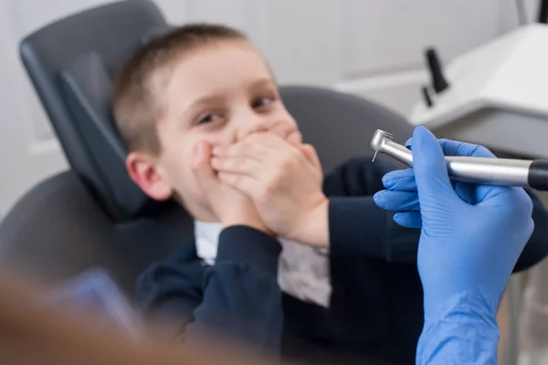 El primer plano del dentista con guantes sosteniendo en la mano broca dental y el niño asustado por los dentistas cubre su boca. La mano dental con broca está en foco — Foto de Stock