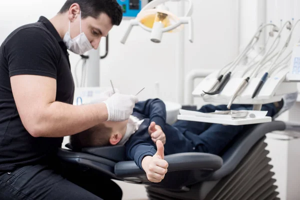 Pediatric tandläkare undersöker tänder av pojken patient i tandvårdsklinik med dental verktyg. Patienten visar tummen. Fokusera på patientens hand. Tandvård — Stockfoto