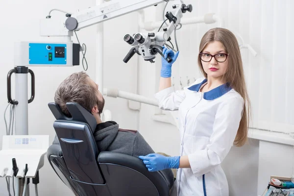 Dentiste féminine attrayante avec microscope se préparer pour la procédure dentaire avec le patient masculin dans le cabinet dentaire moderne. Docteur regardant vers la caméra. La dentisterie. Équipement de stomatologie — Photo