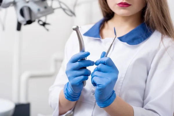 Young woman dentist with blue gloves holding tools - drill and mirror at the dental office. Close-up, selective focus on tools. Dentistry