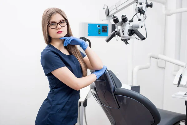 Porträt der schönen Zahnärztin in der modernen Zahnarztpraxis. Arzt mit Brille, blauer Uniform, blauen Handschuhen und Blick in die Kamera. Zahnmedizin — Stockfoto