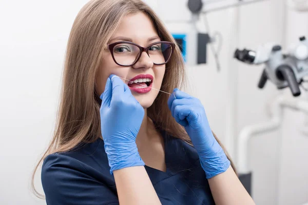 Gros plan de la jeune belle dentiste avec fil dentaire dans la clinique dentaire. Médecin en uniforme bleu et gants, lunettes. La dentisterie. Équipement de stomatologie — Photo