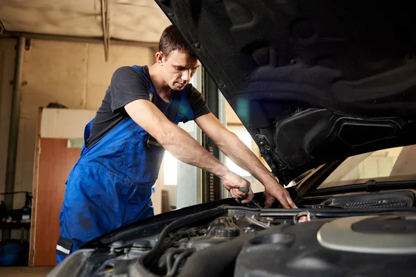 An auto mechanic in a dirty work uniform repairs a car in the garage. The bonnet is open, the guy looks carefully at the problem