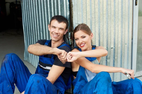 Dos jóvenes mecánicos en trajes azules están contentos después del trabajo y se relajan cerca del servicio de automóviles . —  Fotos de Stock