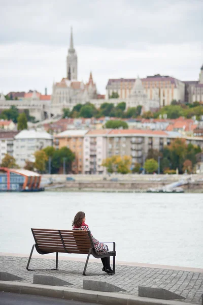 Back view woman sits on a bench on the embankment of Budapest near river and view of Buda side of Budapest with the Buda Castle, St. Matthias and Fisherman's Bastion. Blurred background.