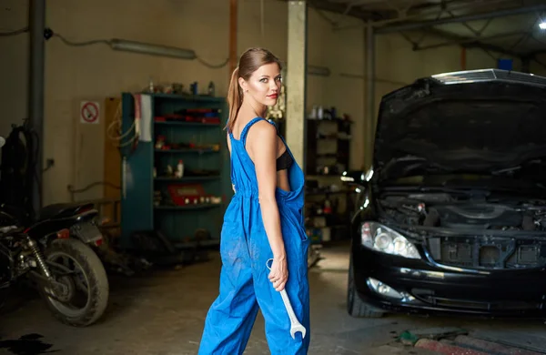 Young female poses with a wrench in her hands in a garage next to the black car and motorbike.