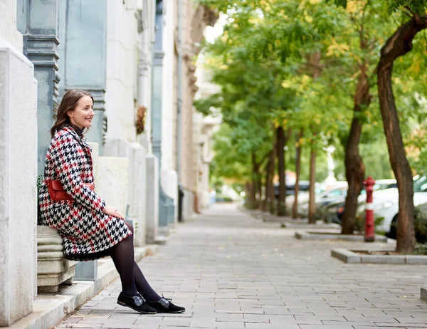 Girl sits near the wall in profile on the autumn street of Budapest. The length of the cave with green trees