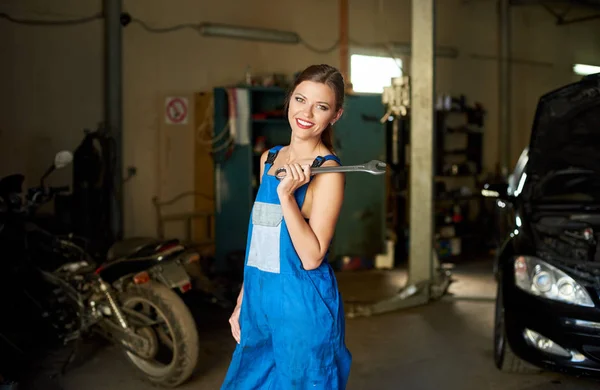 Belle fille mecanicien en vetements de travail dans le garage de reparation. Il tient une cle sur son epaule et regarde la camera avec un sourire Photo de stock par anatoliy gleb