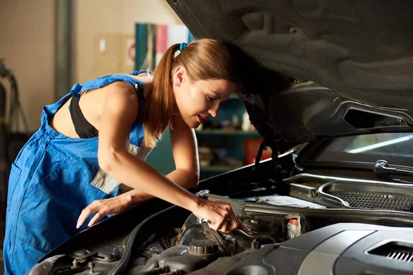 Young car mechanic woman repairing car at service station. Bautiful woman with wrench in her hand, dressed in blue work uniform