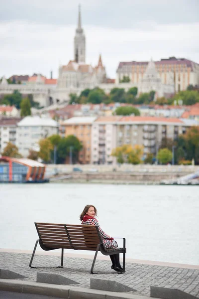 Morena hembra se sienta en un banco en el terraplén de Budapest y se vuelve hacia atrás mirando a la cámara de nuevo la vista de Buda lado de Budapest con el Castillo de Buda, St. Matthias y Bastión del Pescador —  Fotos de Stock