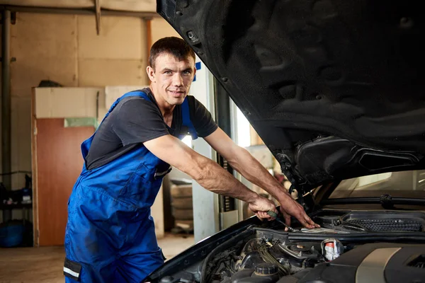 Close-up of mechanic in a dirty work uniform repairs a car in his repair shop. The bonnet is open, the guy looks at the camera