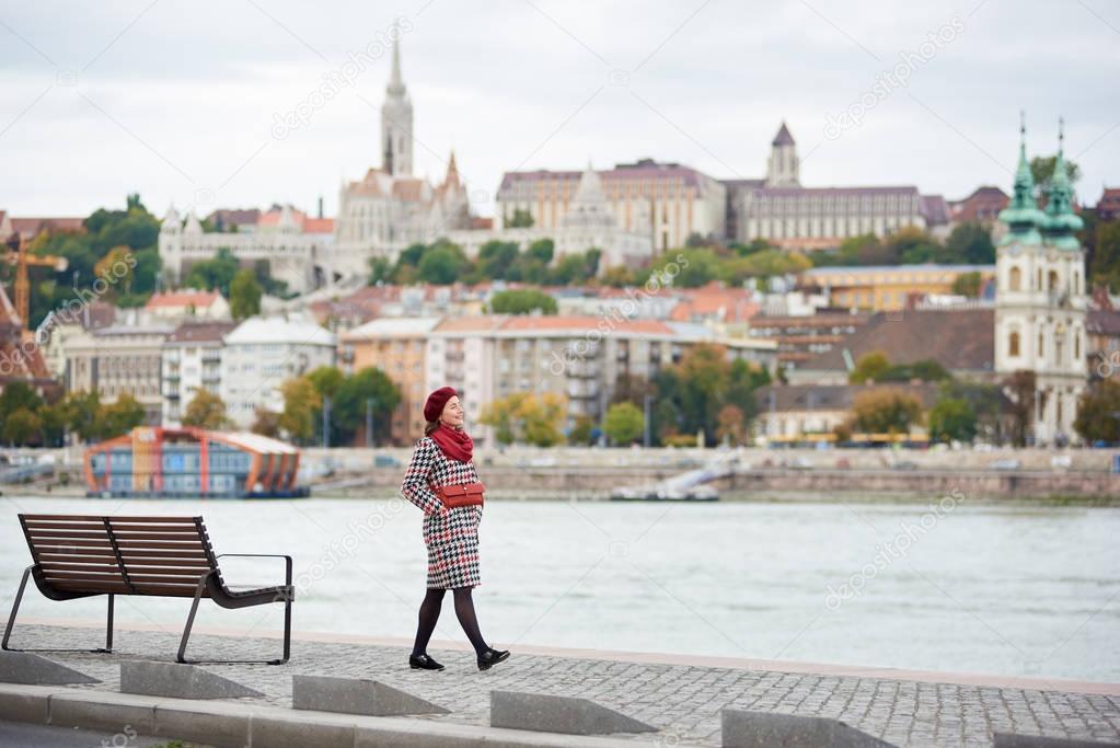 A stylish girl walks along the embankment of Budapest with blurred view of Buda side of Budapest with the Buda Castle, St. Matthias and Fisherman's Bastion