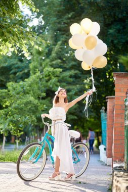 mutlu genç esmer yeşil park sokak üzerinde renkli balonlar ile mavi Bisiklet yakınındaki beyaz elbise sicilini yaz gün 