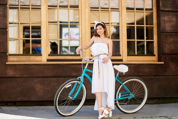 young woman with snow-white smile posing in light dress and bow on head with vintage bicycle on wall with big windows