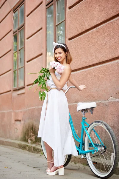 古い壁の背景とレトロな自転車の近くの手で牡丹を保持光のドレスを着た美しい女性 — ストック写真