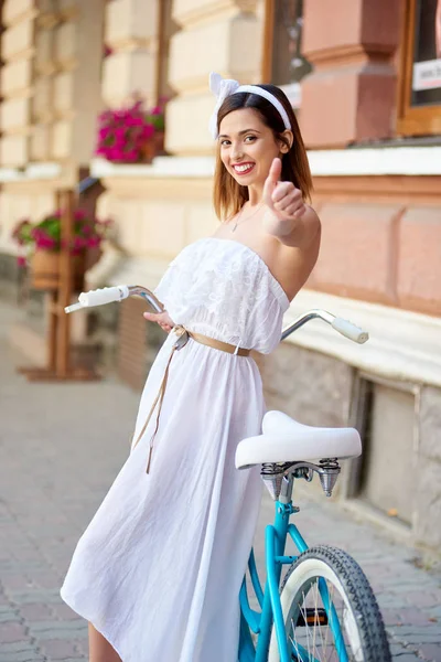 白い夏のかわいい女性のドレスし 旧市街にブルーのレトロな自転車の近くに立っている間親指を出て頭にリボン — ストック写真