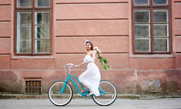 穿着白色连衣裙的美女微笑着捧着鲜花 骑着蓝色的自行车沿着旧的美丽的街道与红色古董建筑背景 — 图库照片