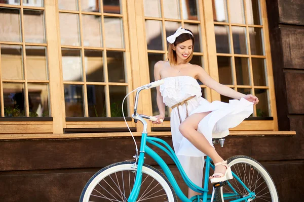 young woman with snow-white smile posing in light dress and bow on head with vintage bicycle on wall with big windows