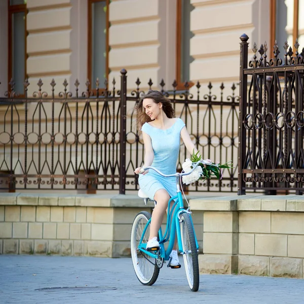 軽い女の子は美しいフェンスと古い建物の背景に彼女の手で牡丹とヴィンテージ自転車に乗ってください — ストック写真