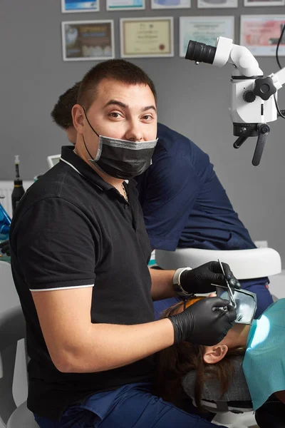 Młodego dentysty w czarnej maski i rękawiczki traktuje pacjenta za pomocą grodzy i mikroskopu. Współczesna Stomatologia z wykorzystaniem nowych technologii. Certyfikaty są widoczne w tle ściany — Zdjęcie stockowe