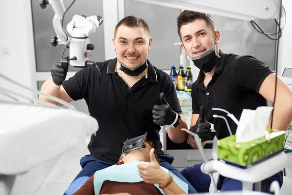 两个穿黑色制服的男牙医在白色牙科办公室坐着 竖起大拇指 微笑着拍照 — 图库照片