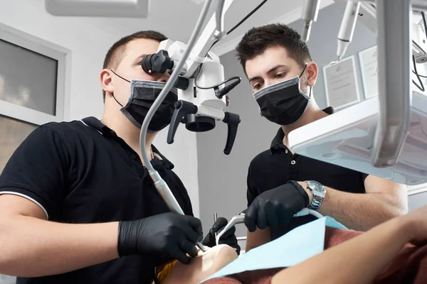Αρσενικό οδοντίατρος φαίνεται στο μικροσκόπιο τη θεραπεία των δοντιών του ασθενούς, ένα άλλο αρσενικό ο γιατρός τον βοηθά — Φωτογραφία Αρχείου