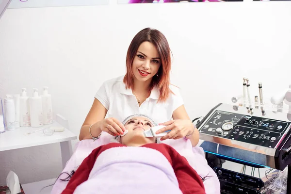 Улыбающийся врач с ультразвуковым скрабером проводит процедуру ультразвуковой очистки лица для женщины. Современная косметологическая клиника . — стоковое фото