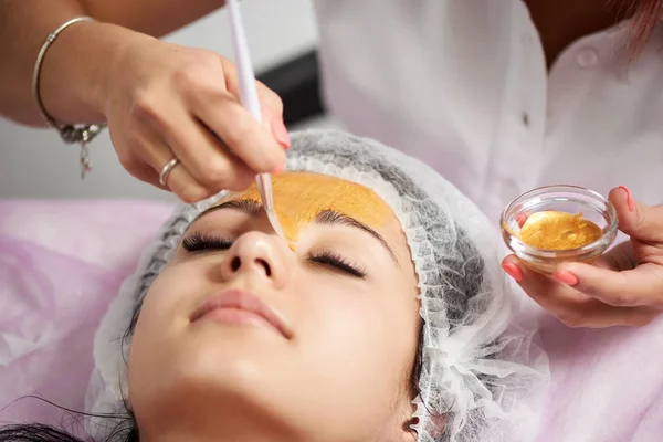 Kvinnliga kosmetolog tillämpar guld mask till en ung kvinna i en skönhetssalong. Kosmetika och hudvård rutin. — Stockfoto