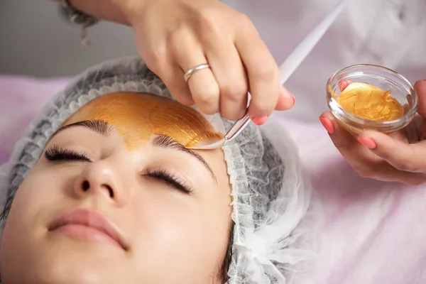 Primer plano del retrato de una hermosa chica con un sombrero especial en la cabeza aplicando una máscara facial dorada. Cosmetología del hardware . — Foto de Stock